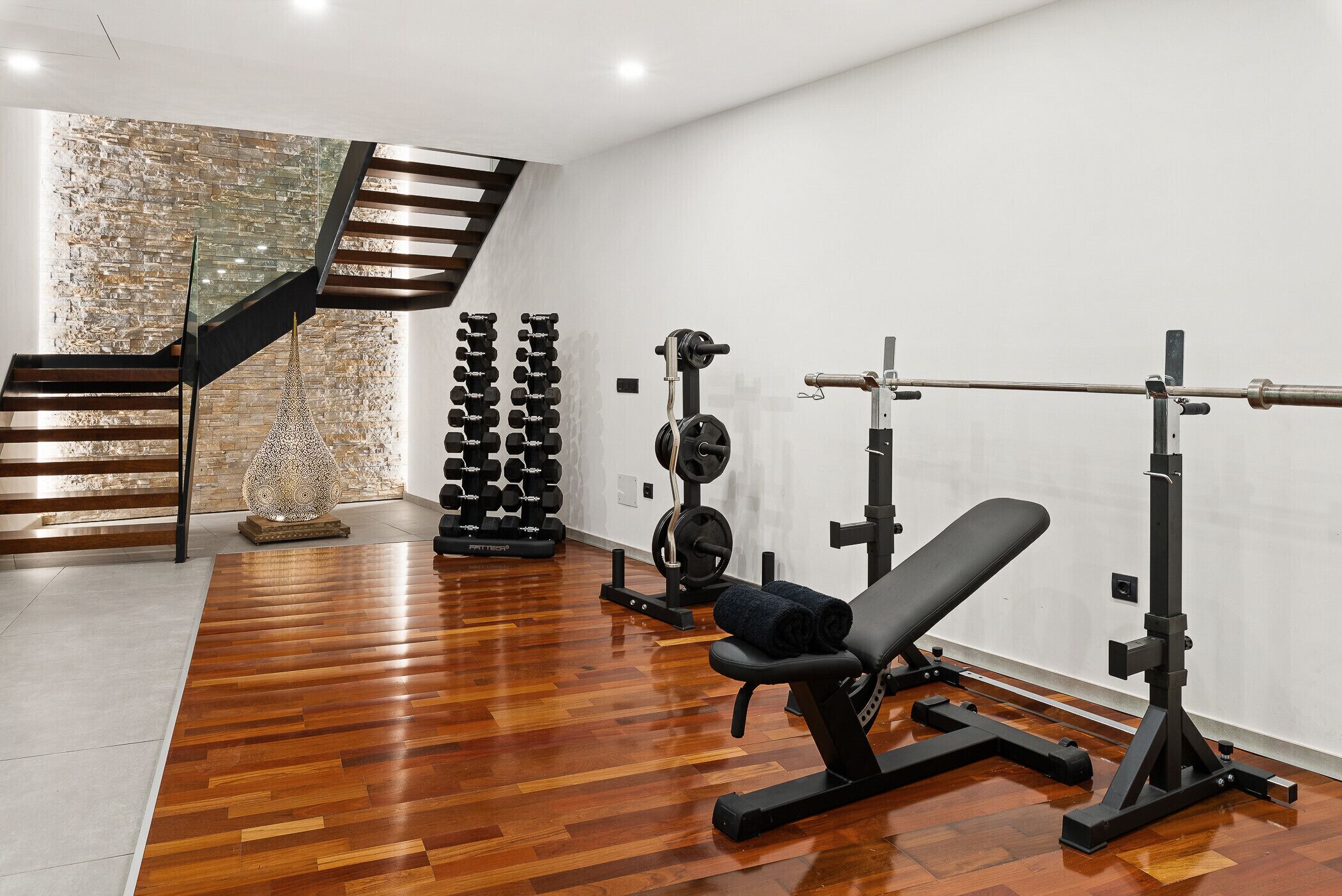 A gym with wooden flooring, a bench press, dumbbells, and a staircase.