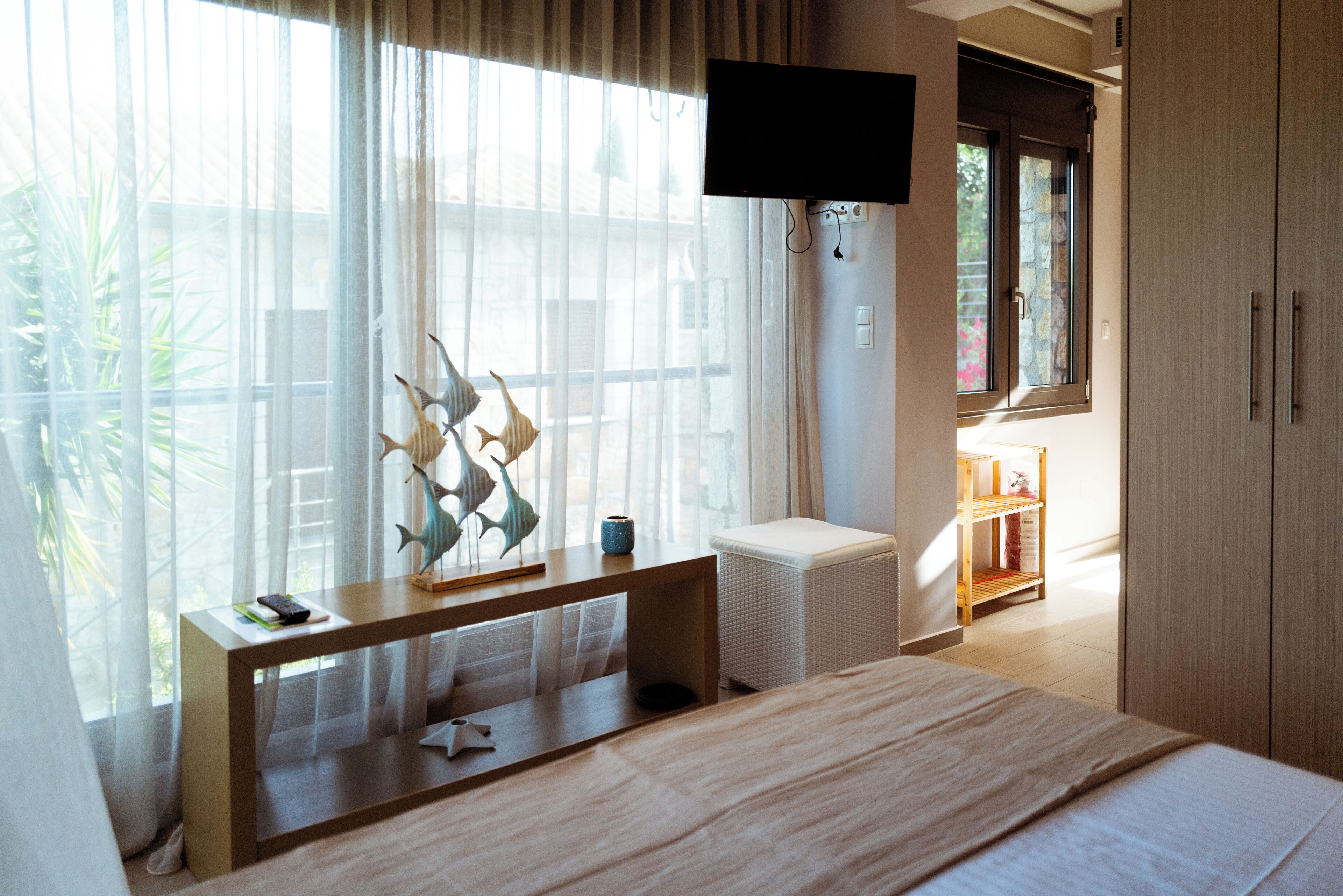 A hotel room with a bed, a TV mounted on the wall, a window with sheer curtains, and a small table with decorative items.
