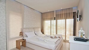 A modern hotel room with a large bed, a small desk, and a large window with curtains. - Christie's Luxury Suites (Igoumenitsa)