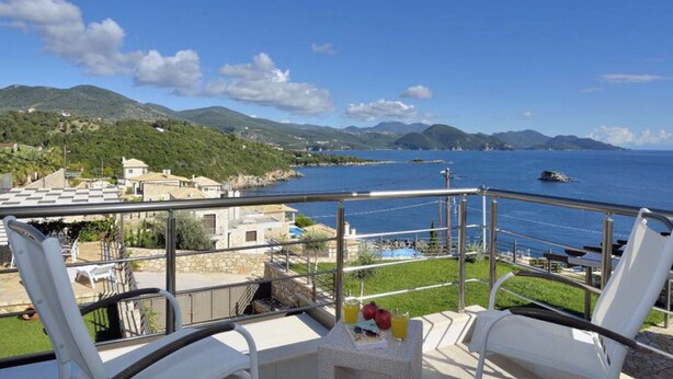 A balcony with white chairs and a table, overlooking a coastal area with houses and a clear blue sky. - Christie's Luxury Suites (Igoumenitsa)