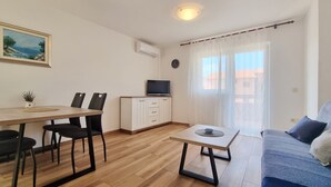 Ein Wohnzimmer mit einer Couch, einem Couchtisch, einem Fernseher auf einem Schrank, einem Esstisch mit Stühlen und einem Gemälde an der Wand.
