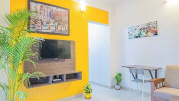 Una sala de estar moderna con una pared de acento amarilla, un televisor montado, una mesa de comedor con sillas y plantas en macetas.