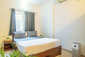 A bedroom with a bed, a bedside table with a plant, a window with curtains, and a standing heater. - Housr Ferguson Apartment Near Radisson (Bengaluru)