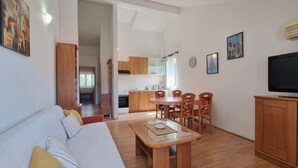 Una sala moderna con un sofá, una mesa de comedor de madera y sillas. Hay una zona de cocina con armarios y una estufa. Un televisor está montado en la pared.
