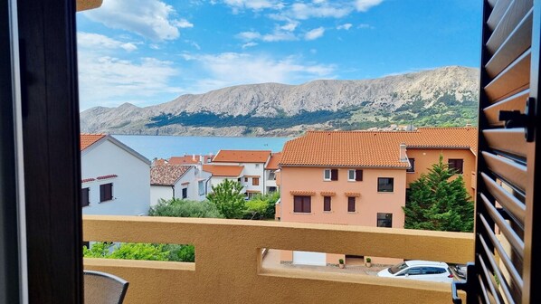 A view from a balcony overlooking a coastal town with terracotta-roofed buildings, a mountain in the background, and a body of water.