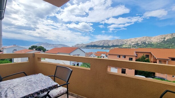 A balcony with a table and chair, overlooking a coastal town with terracotta-roofed buildings and a mountainous backdrop.