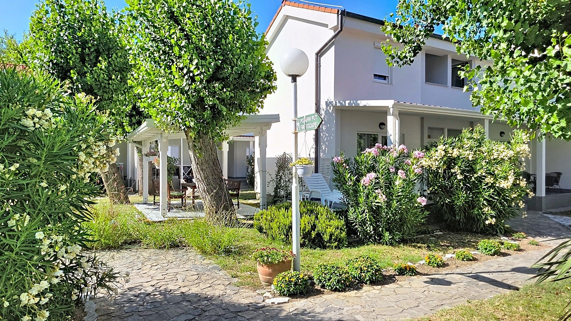En boligbygning med et overdækket udendørs siddeområde, skilt med brandstige og en velholdt have med forskellige planter og blomster.