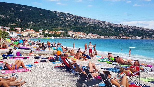 A crowded beach with people sunbathing and swimming, surrounded by buildings and hills.