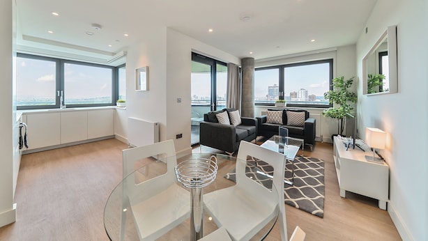 A modern living room with a glass dining table, black and white sofas, and large windows offering city views.