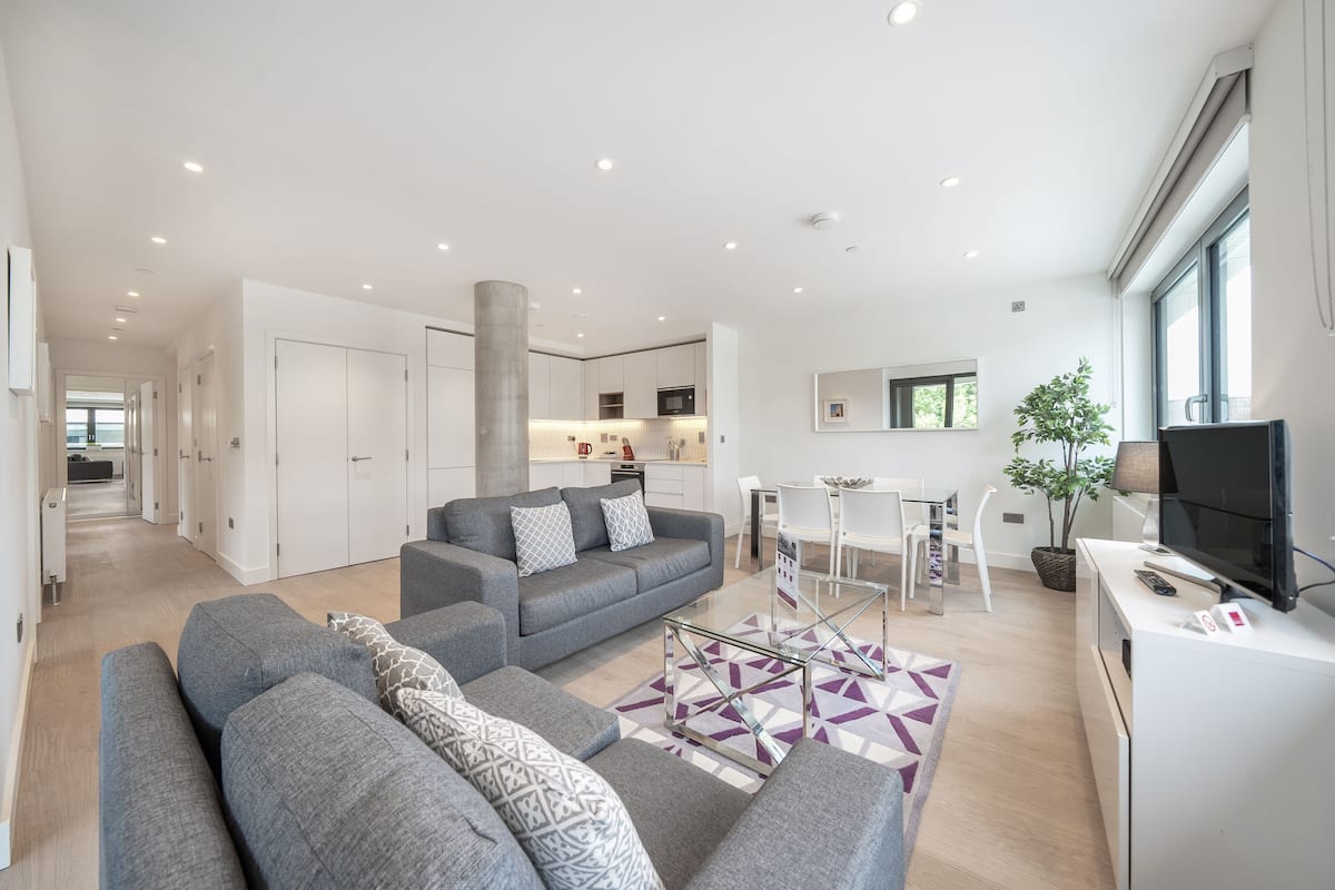A modern living room with a grey sofa set, a coffee table, and a TV. There is a kitchen in the background with white cabinets and a dining area with a table and chairs.