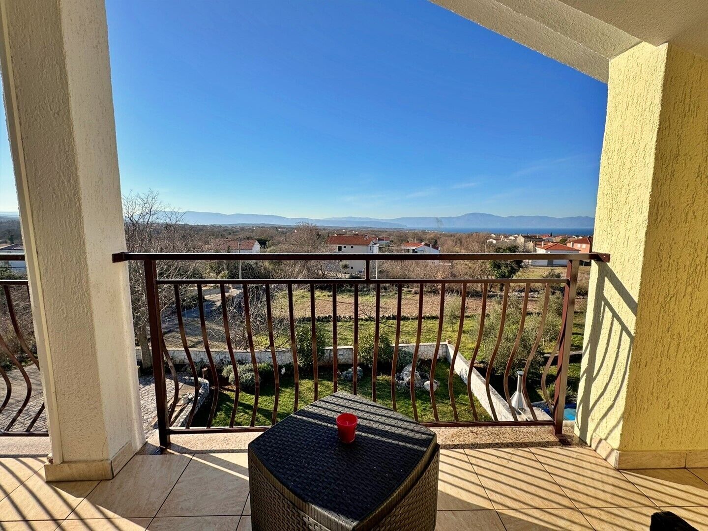 A balcony with a wicker table and a red cup, overlooking a residential area with houses and a distant mountain range.