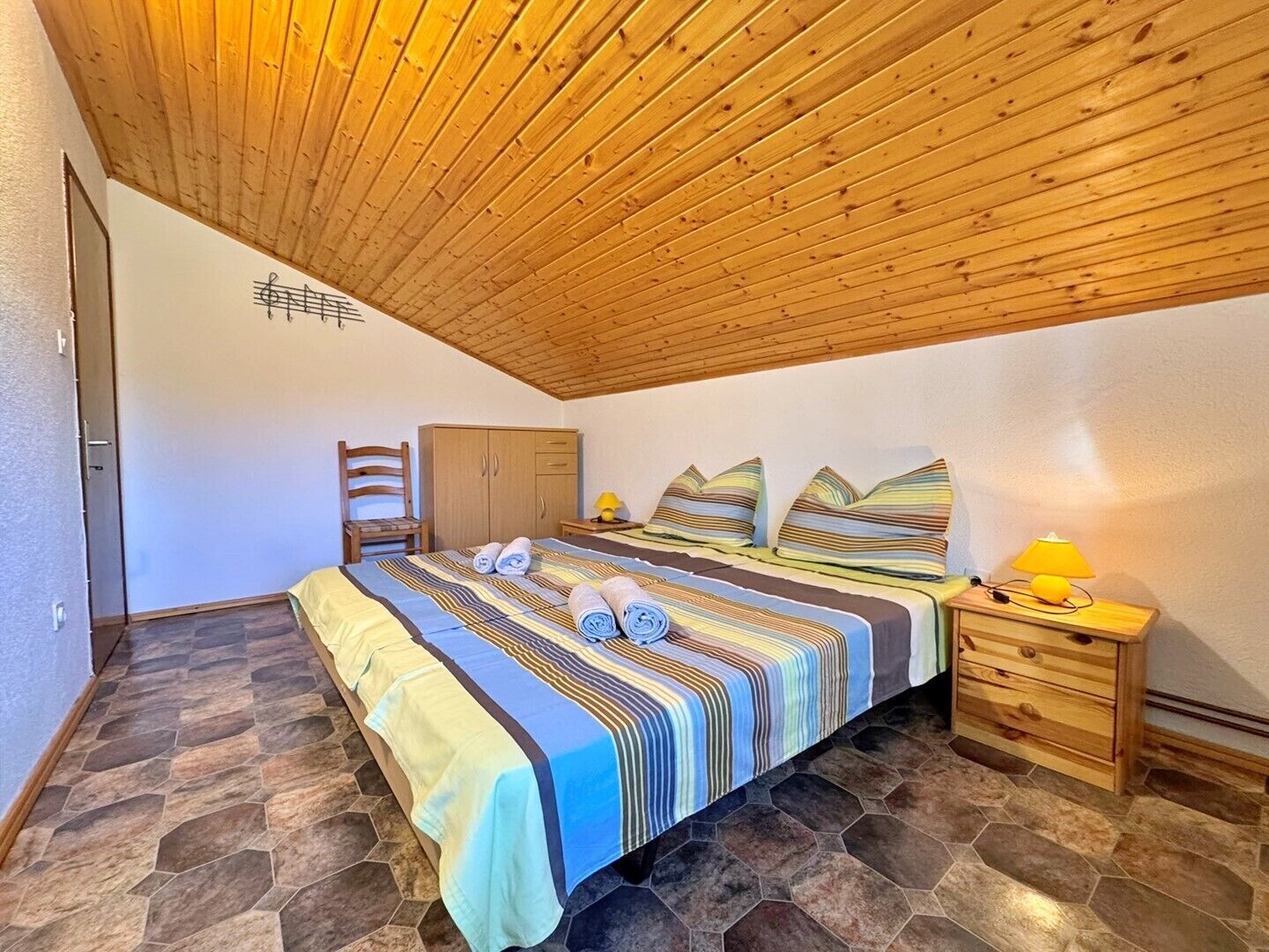 Un dormitorio con techo de madera, una cama con ropa de cama a rayas, una silla, un armario y una mesita de noche con una lámpara.