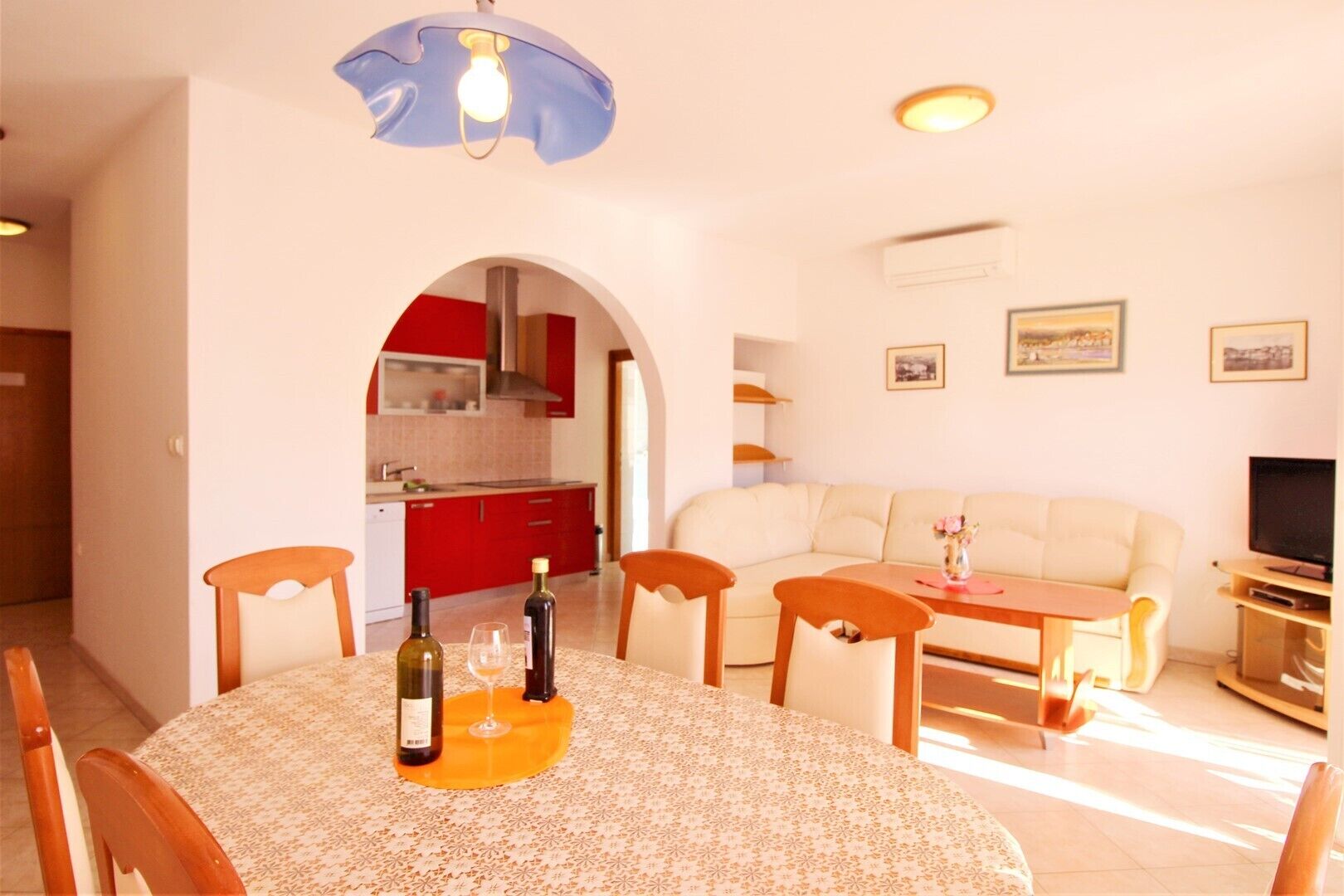 A dining area with a round table, chairs, and a bottle of wine. There is a kitchen visible through an archway, a sofa, and a television in the living area.