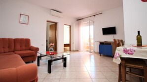 A living room with a sofa, a coffee table with a vase, a dining area with a table and chairs, a television, and a kitchen area. - Apartment Lucijana No.2 - with Balcony, 150 m from the Beach (Baska)