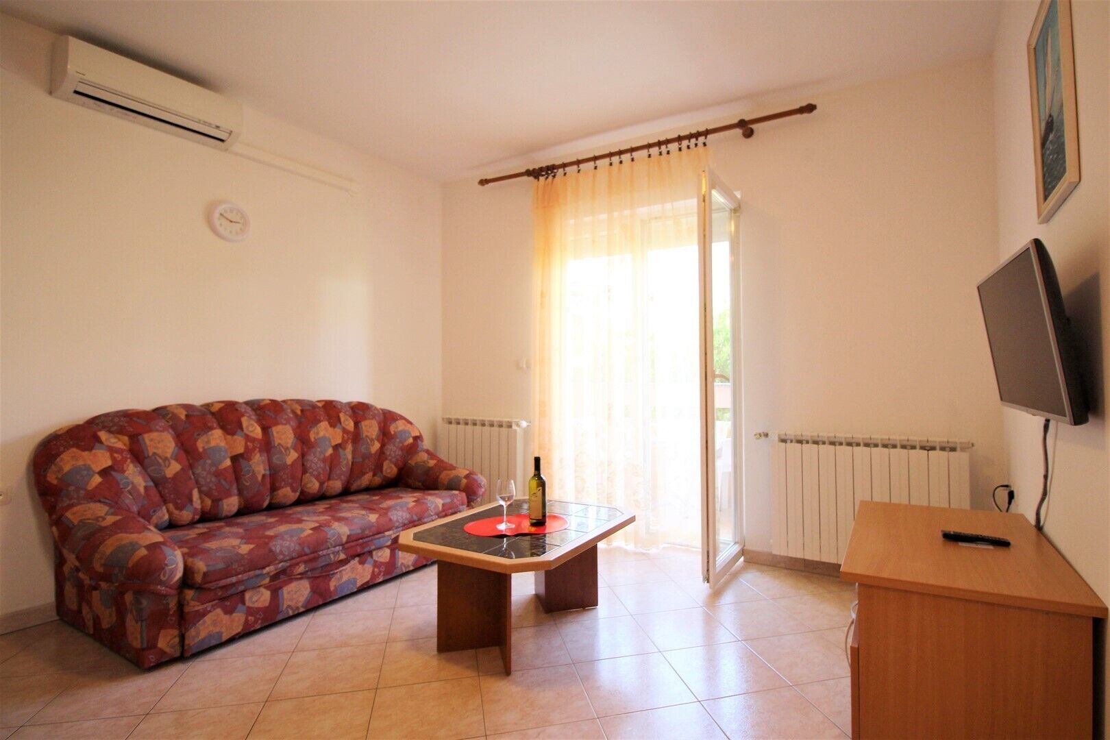 A living room with a patterned sofa, a small wooden table with a bottle and glass, a TV on a stand, and a window with curtains.
