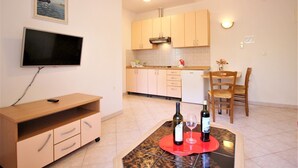 Un espacio habitable compacto con un televisor montado en la pared, un mueble bajo para la televisión de madera, una zona de comedor con mesa y sillas, y una cocina con armarios y estufa.