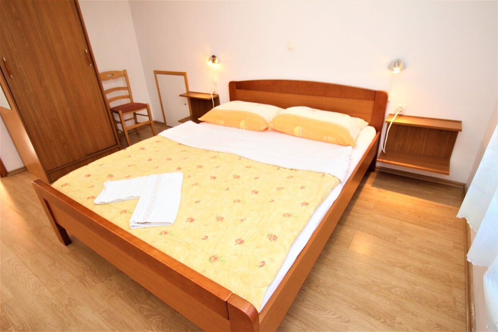 Ein ordentlich bezogenes Bett mit floralem Überwurf, zwei Nachttische mit Lampen, ein Holzstuhl und ein Holzschrank.