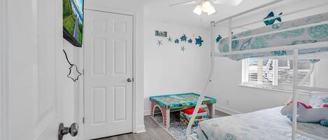 A child's bedroom with bunk beds, a ceiling fan, a small table, and a window with blinds.