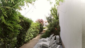 A narrow pathway flanked by dense foliage and a white wall, leading to a seating area with white plastic chairs and a table. - Studio Mul No.4 Bs - for 4 Persons, with Terrace (Baska)