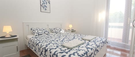 A bedroom with a large bed, two bedside tables with lamps, a framed artwork on the wall, and a view of the outdoors through a window.