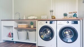 A laundry area with two white washing machines, a wooden shelf with storage bins, a small plant, and a sign indicating laundry hours.