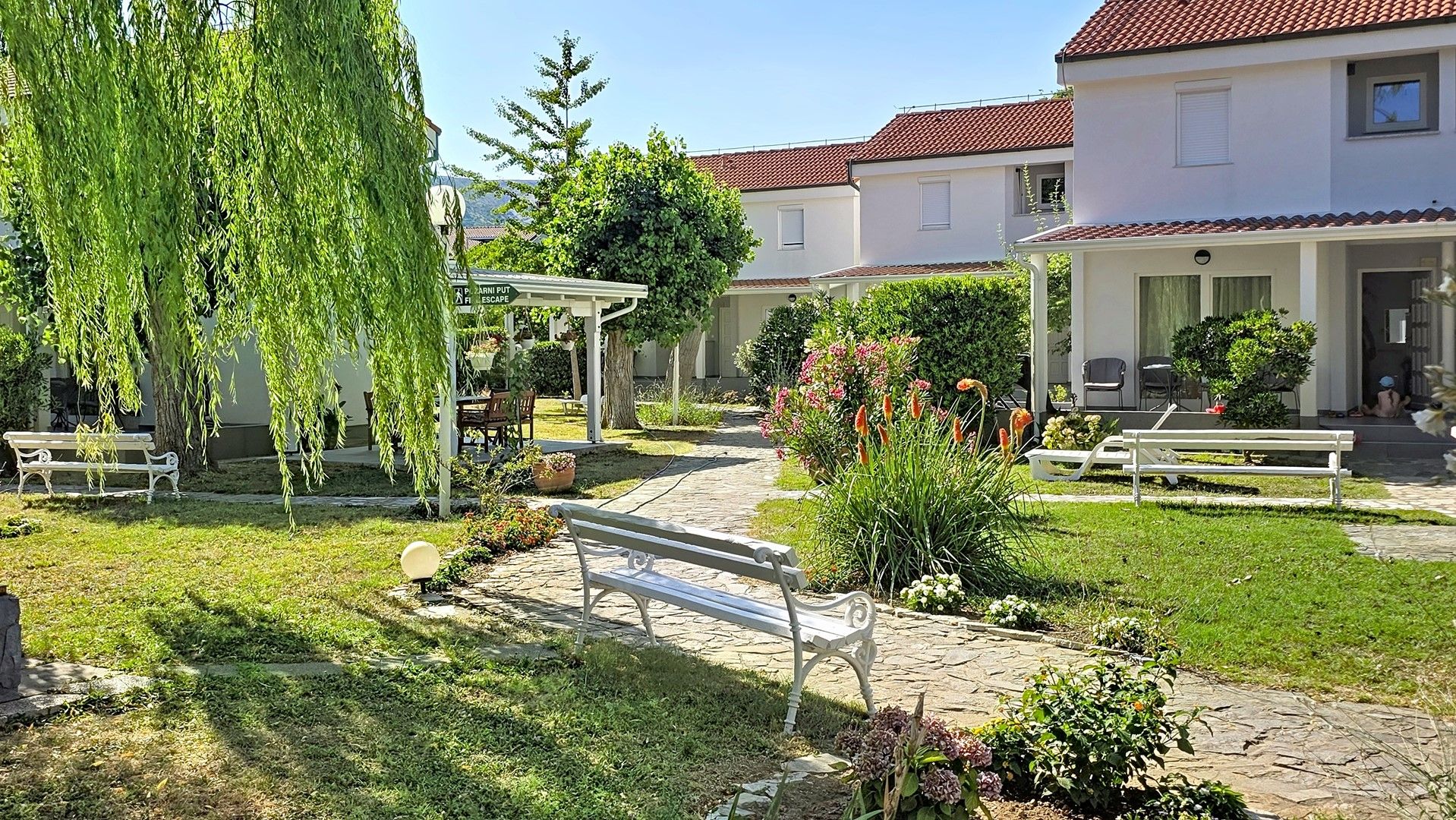 A courtyard with a bench, a pergola, and a pathway leading to a building with a red-tiled roof.