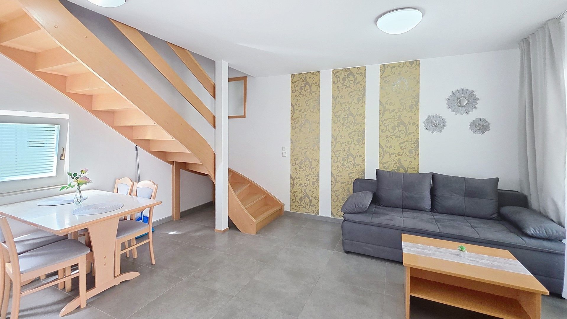 Een moderne woonkamer met een grijze bank, een houten trap en een eethoek met een tafel en stoelen.
