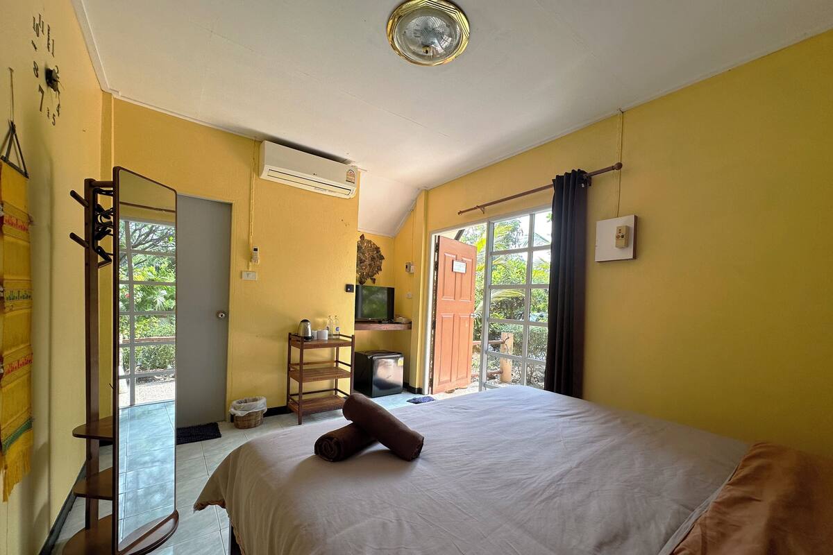 A bedroom with a bed, a TV, a small table with a kettle, and a window with curtains.