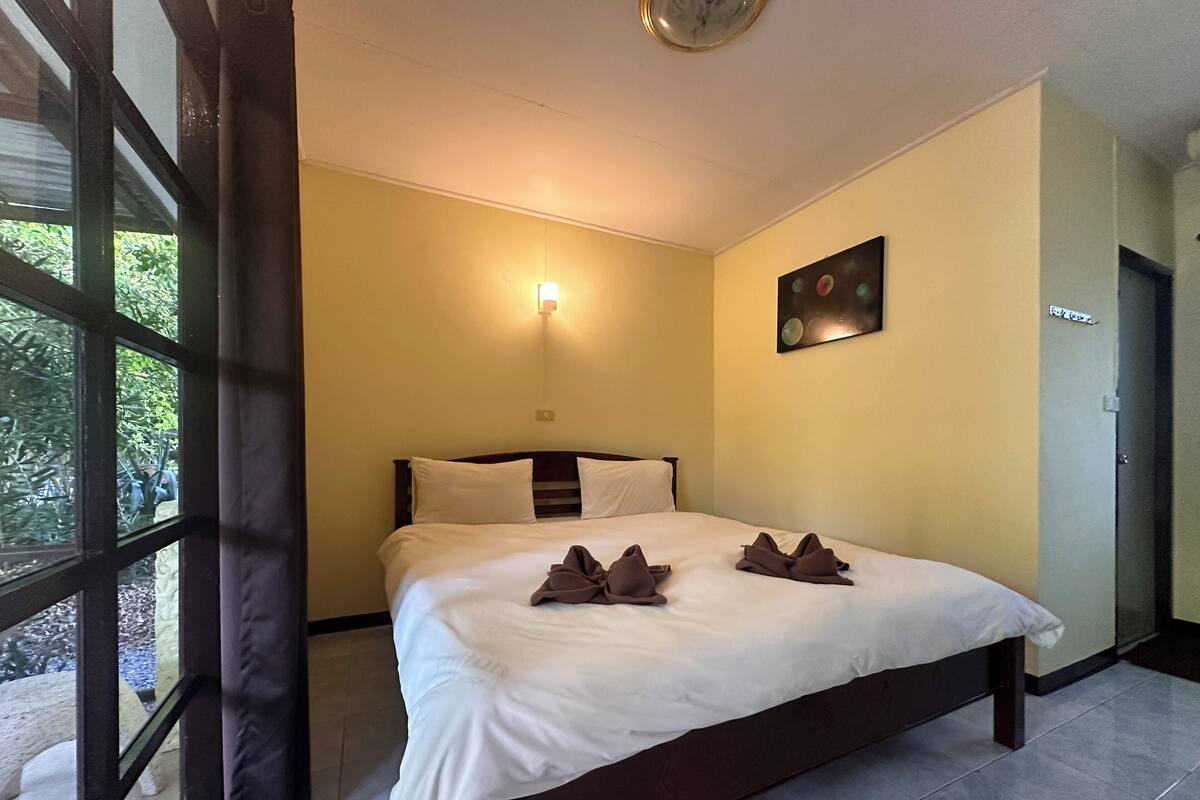 A hotel room with a bed, a wall-mounted light, a painting, and a window with curtains.