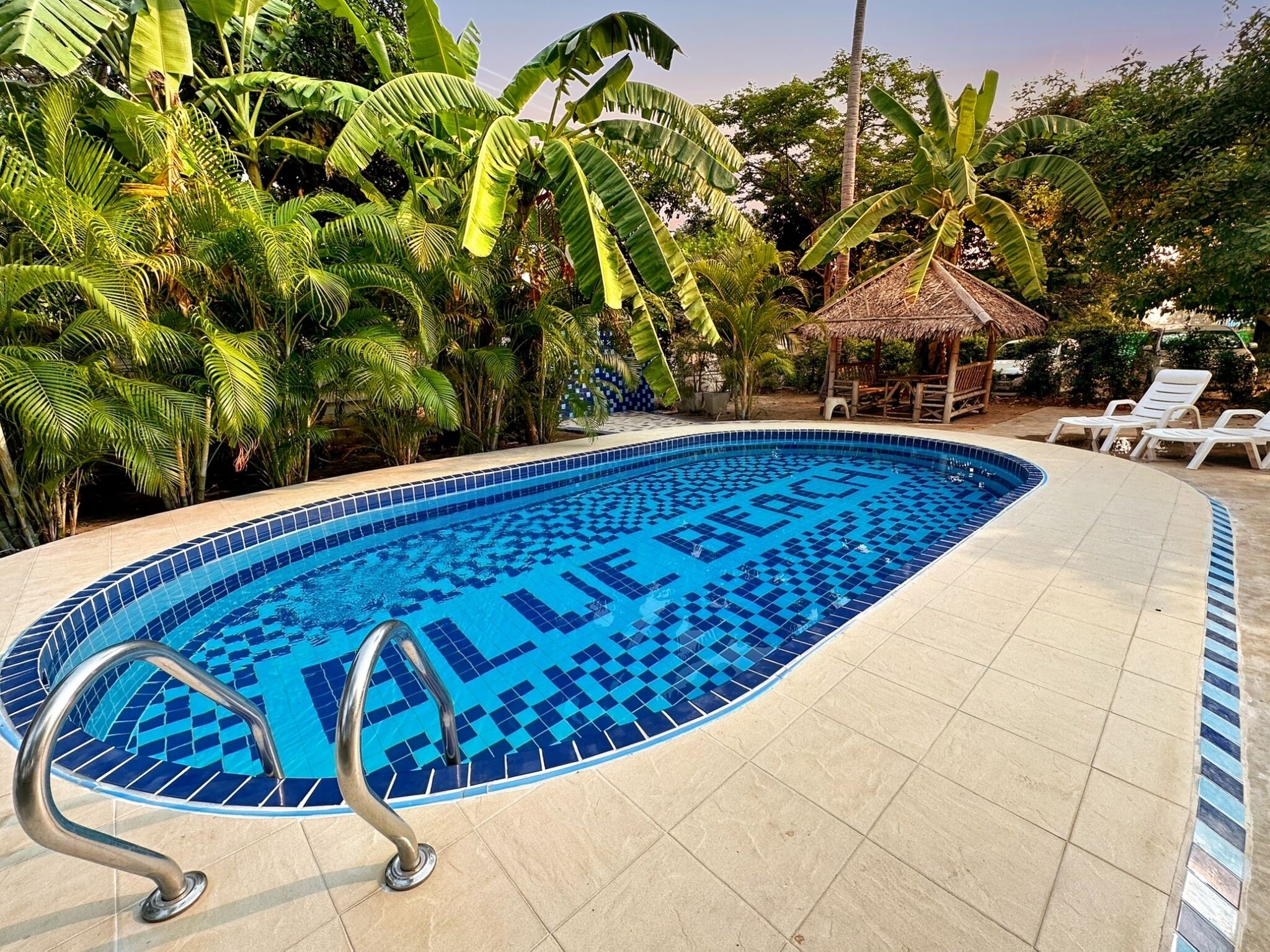 Una piscina con un diseño azul de mosaicos, rodeada de abundante vegetación y una estructura de techo de paja.