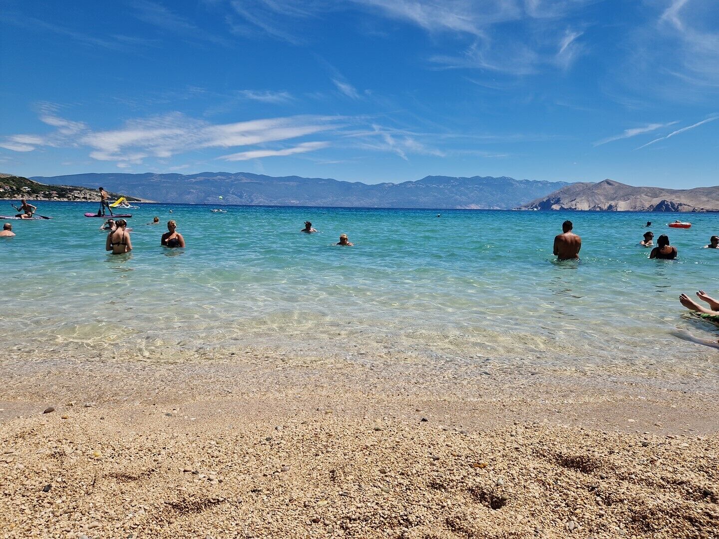 A beach with clear turquoise water, pebbly shore, and distant mountains.