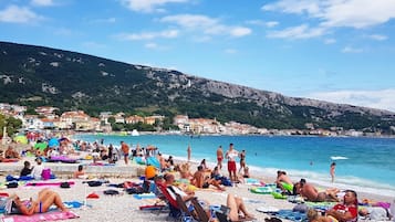 Una playa concurrida con gente tomando el sol y nadando, rodeada de edificios y colinas.