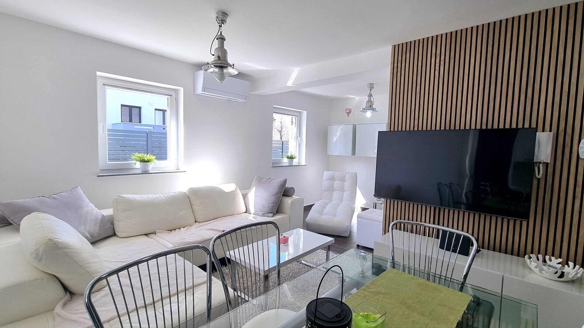 A modern living room with a sofa, chairs, a glass coffee table, and a TV mounted on a wooden panel.