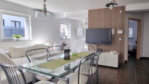 A modern living room with a glass dining table, white sofas, and a TV mounted on a wooden panel wall.