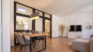 Ein modernes Wohnzimmer mit einem Holzes Esstisch, grau gepolsterten Stühlen, einem grauen Sofa, einem Flachbildfernseher und einem weißen Schrank.