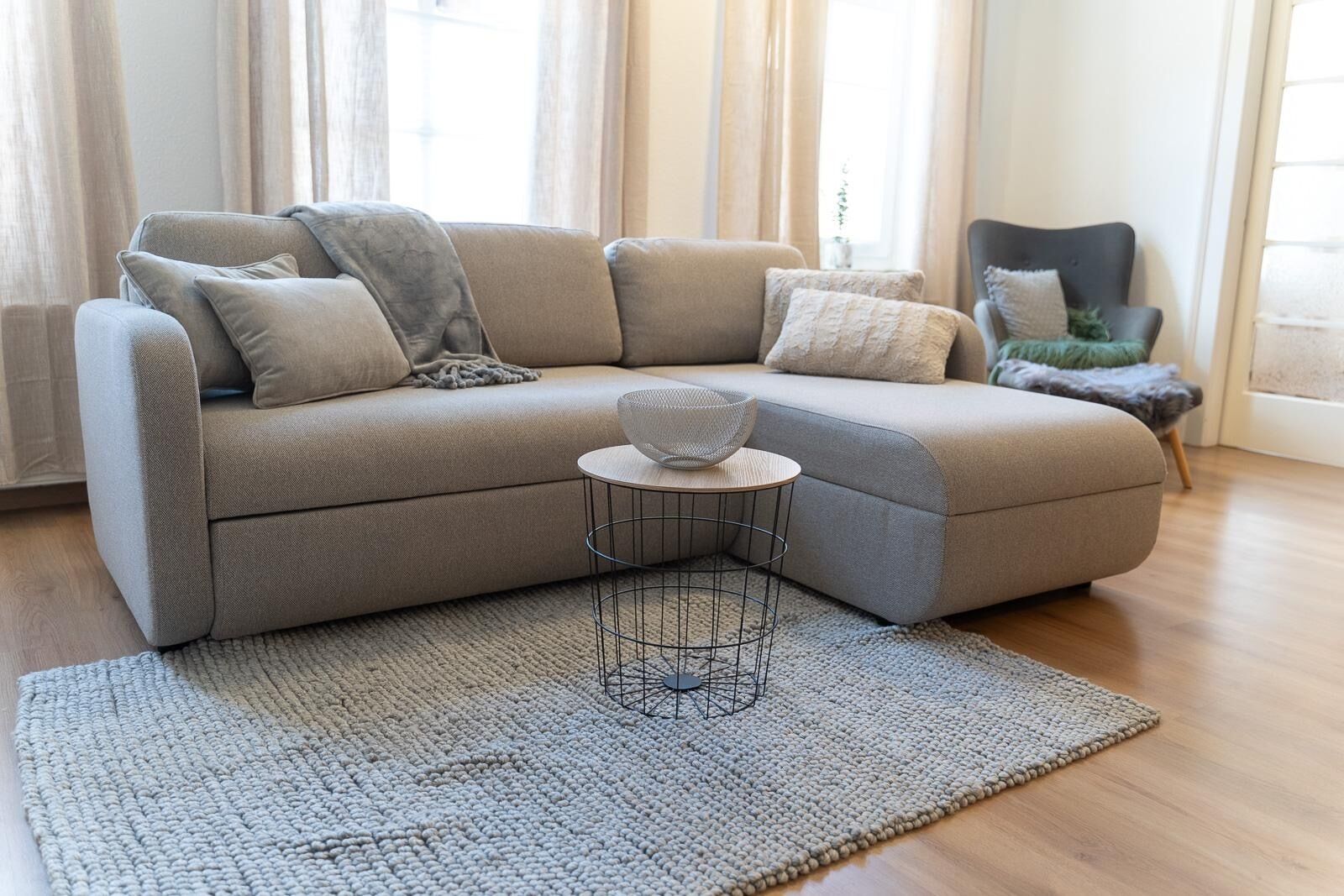 Un salon moderne avec un canapé d’angle beige, une table basse ronde surmontée d’un bol en fibres tressées et un tapis à motifs.