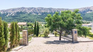 Una entrada empedrada con pilares de piedra, una reja de metal y vistas de un paisaje montañoso con vegetación y edificios al fondo.
