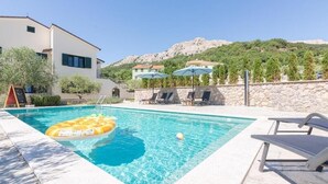 Une piscine avec des chaises longues et des parasols, entourée d’un mur de pierre et avec une chaîne de montagnes en arrière-plan.