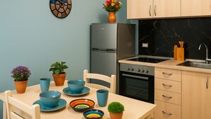 A modern kitchen with a refrigerator, stove, and sink. There is a dining table with chairs and various dishes and plants.