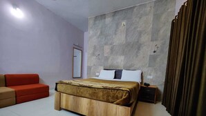 A bedroom with a bed, a wooden nightstand, a red sofa, and a large window with curtains. - Aditya Forest Eco Resort (Abu Road)