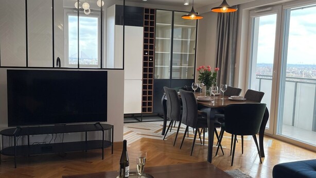 Una sala de estar moderna con una televisión de pantalla plana, una mesa de centro con una botella y un vaso, un sofá, un comedor con mesa y sillas, y grandes ventanas con vistas a la ciudad.