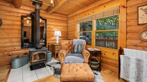 Een gezellig blokhutinterieur met een houtkachel, een comfortabele fauteuil, een bank en een groot raam met uitzicht op de natuur.