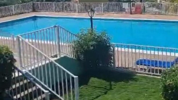 En swimmingpool med udsigt til havet, omgivet af et hegn og grønt vegetation.
