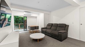 Un salon moderne avec un canapé gris, une table basse ronde et une petite table à manger avec des chaises. Une porte-fenêtre coulissante donne accès à un balcon agrémenté de plantes.