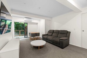 A modern living room with a grey sofa, a round coffee table, and a small dining table with chairs. There is a sliding glass door leading to a balcony with greenery outside.