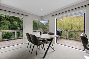 A modern dining area with a large table and chairs, large windows with a view of greenery, and a balcony with a barbecue grill.