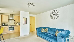 A modern living room with a blue sofa, a yellow chair, a kitchen in the background, and a wall clock. - Charming 2-Bedroom Flat in South Croydon (Greater London)