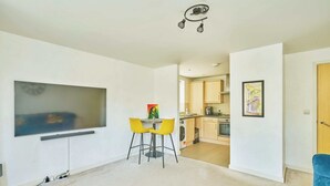 A modern apartment with a flat-screen TV, a dining area with yellow chairs, a kitchen with wooden cabinets, and a blue chair in the living room. - Charming 2-Bedroom Flat in South Croydon (Greater London)