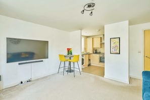 A modern apartment with a flat-screen TV, a dining area with yellow chairs, a kitchen with wooden cabinets, and a blue chair in the living room.