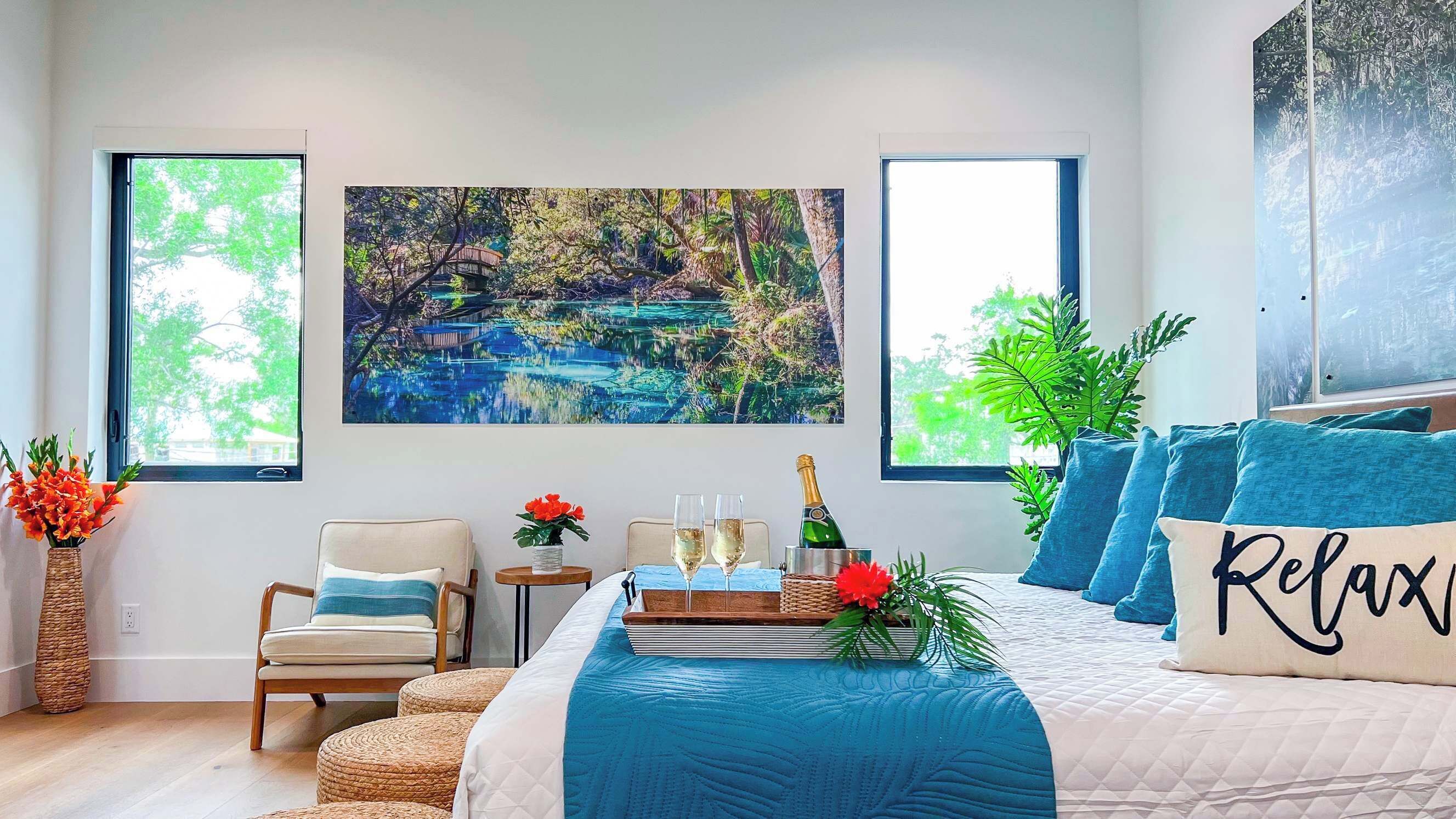 Um quarto com uma cama grande, uma mesinha com copos e uma garrafa, e um quadro de uma paisagem.
